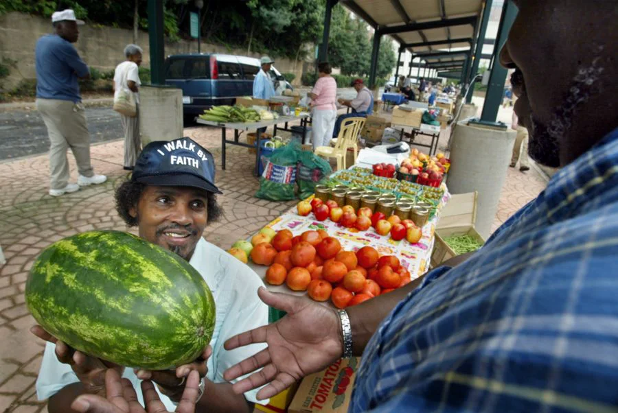Vendor at a farmers market holding a watermelon and speaking with a customer at a produce stand.