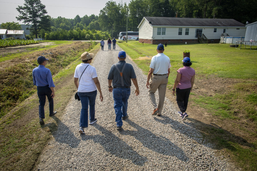 A white Holiday tour bus stuck in sandy ground, with deep tire ruts in front of it. Several people stand around the scene on the open field, some observing and one gesturing toward the bus. Pickup trucks are parked in the background near a treeline under a partly cloudy sky.