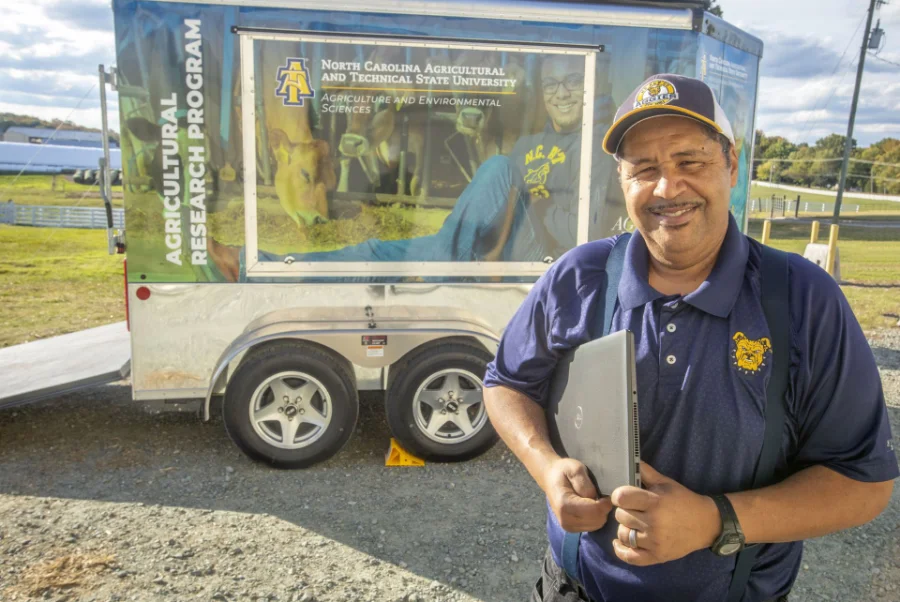 A smiling man holding a closed laptop stands outside in front of an Agricultural Research Program trailer featuring a North Carolina Agricultural and Technical State University banner.