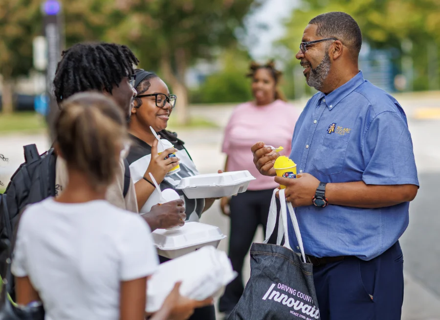 A smiling man wearing glasses and a blue button-down shirt stands outside talking with a group of students holding takeout food containers and frozen treats.