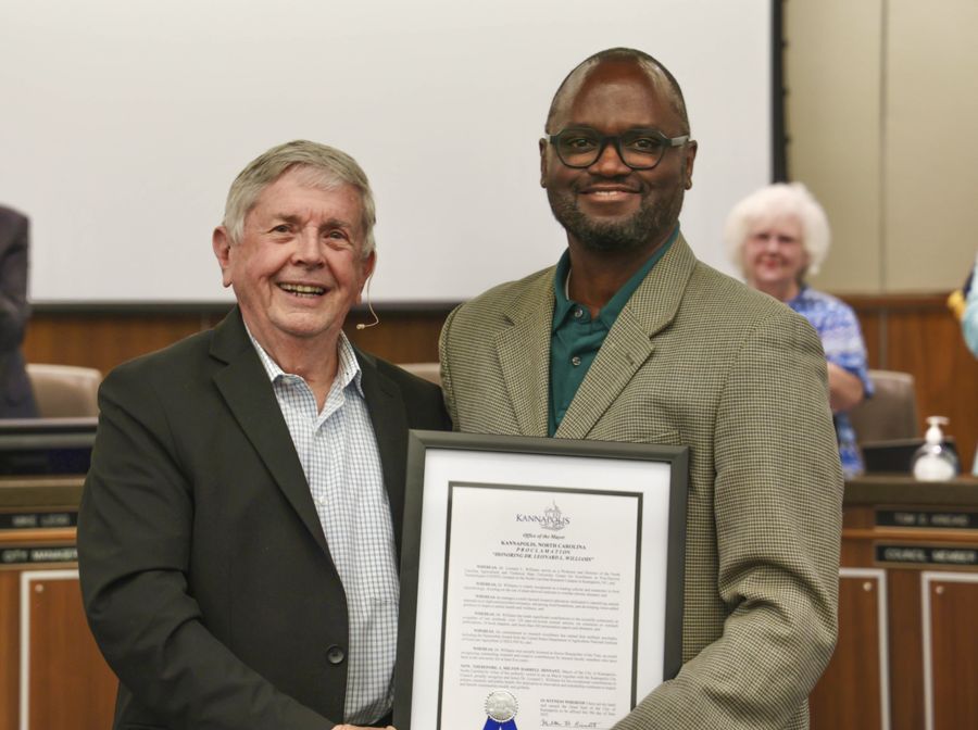 Two men stand together smiling; one holds a framed certificate. In the background, people applaud in a formal setting with a wood-paneled wall and a large screen.