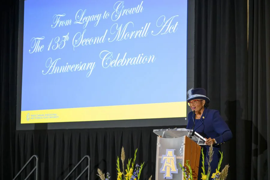 A woman in a blue suit and matching hat speaks at a clear podium on a stage. Behind her is a large screen displaying the text “From Legacy to Growth: The 133rd Second Morrill Act Anniversary Celebration.” Floral arrangements sit in front of the podium, and dark curtains frame the scene.