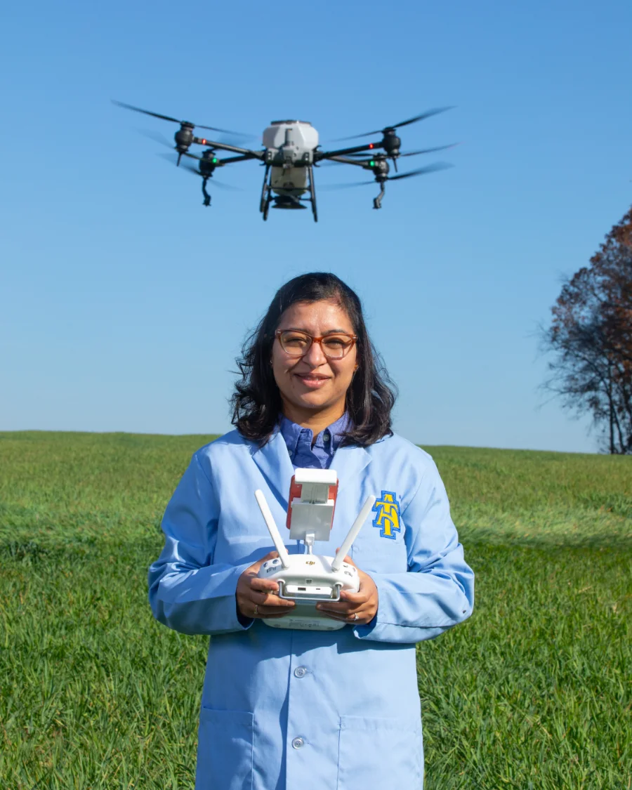 A woman in a blue lab coat stands in a green field operating a drone, which hovers in the air above her. Text explains N.C. A&T’s agricultural innovation, including work with climate-smart soil, specialty crops, and food as medicine.