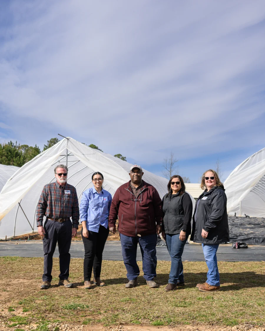 Five adults stand outdoors in front of white agricultural tents, representing North Carolina’s small farmers, STEM educators, and community leaders. The text highlights N.C. A&T’s efforts to empower these groups through Cooperative Extension work.