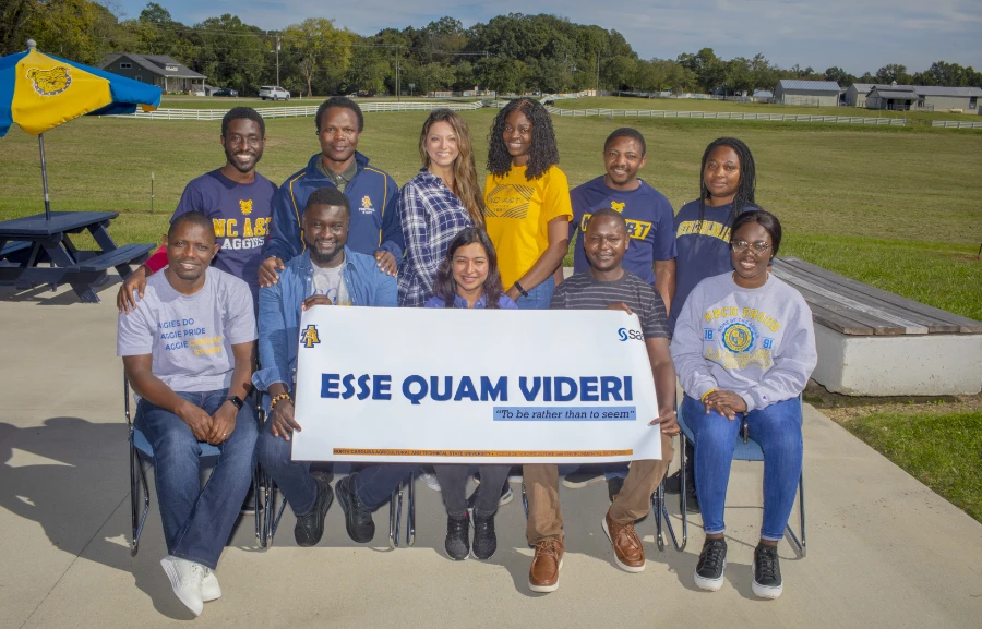 A group of smiling people outdoors, seated and standing behind a banner reading "Esse Quam Videri - To be rather than to seem," with a grassy field and buildings in the background.