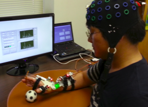 A participant wearing a multi-electrode EEG cap sits at a desk, focusing on a small black-and-white soccer ball. Their right hand is fitted with a motorized, white hand orthosis device equipped with small red indicator lights and attached wiring. In the background, computer monitors display real-time signal graphs and software interfaces. This experimental setup demonstrates the initial assessment of a Brain-Computer Interface (BCI)-driven hand orthosis, designed to evaluate its wearing comfort, usability, and potential efficacy for the motor rehabilitation of stroke patients.