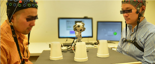 Two participants are sitting across a table from one another, both wearing multi-electrode EEG caps to record brain activity for a collaborative Brain-Computer Interface (BCI) experiment. In the center of the table between them is the "NC State Brainbot," a small robotic arm holding a miniature soccer ball, positioned behind three inverted white cups. In the background, two computer monitors display the BCI system's software interfaces. The participants are focused on the task, and their eyes have been anonymized with black rectangles.