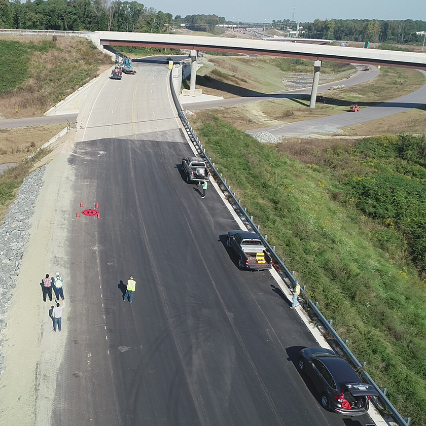 A photo of an inspection team standing on a road near a bridge sending a drone for inspection. A drone landing pad is nearby.