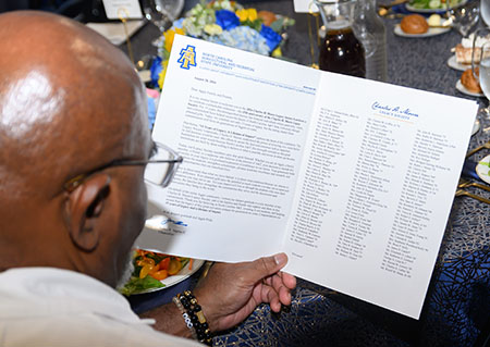 The back of the head of a bald man wearing glasses looking at the event program on a table surrounded by plates of food